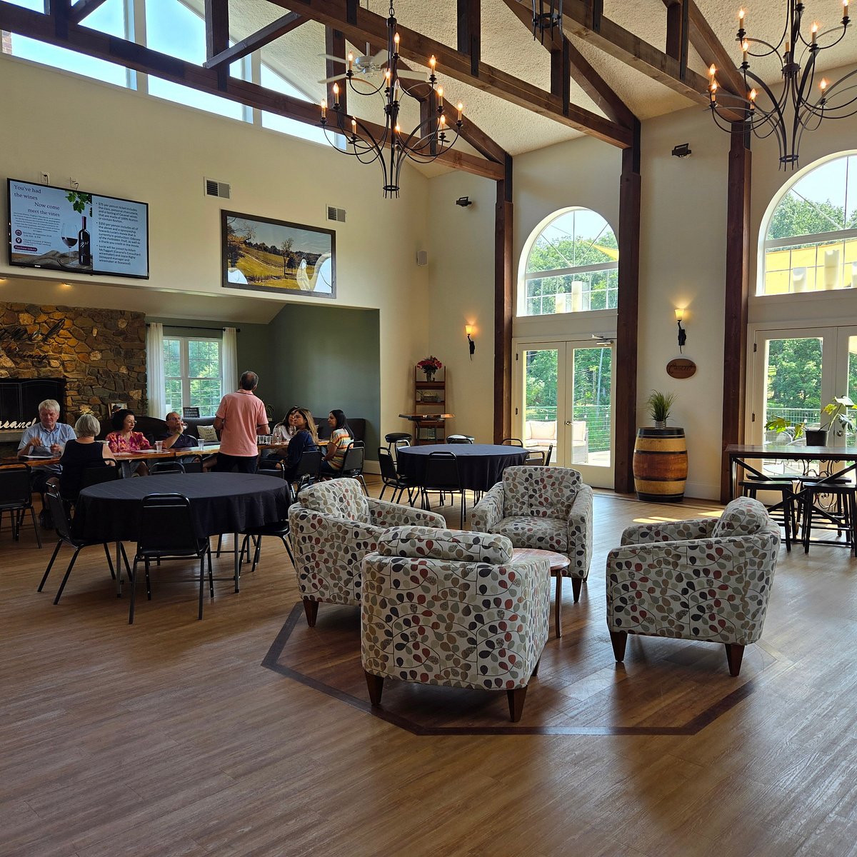 High-ceilinged tasting room with timber beams, chandeliers, a stone fireplace, round tables and armchairs